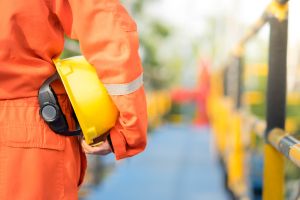 Un ouvrier de dos, en combinaison de travail orange, tenant un casque de chantier jaune sous le bras, marchant sur une plateforme industrielle.
