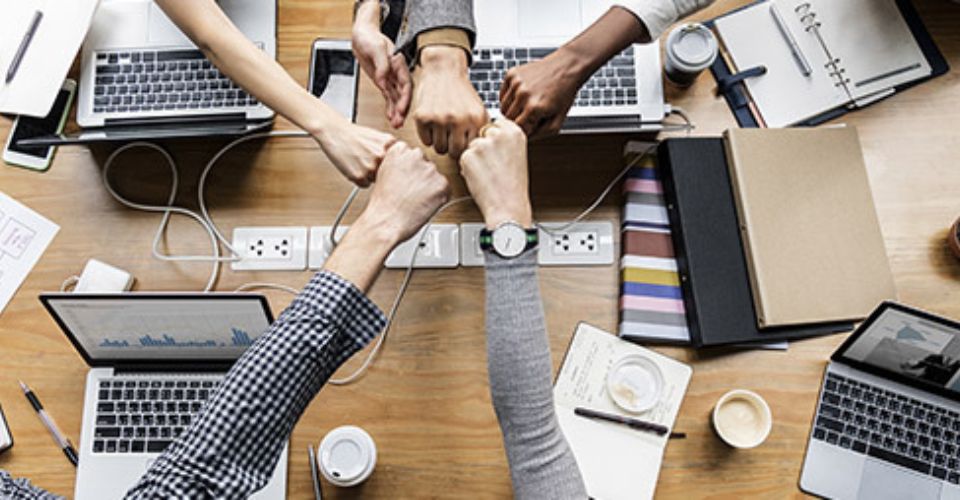 Plusieurs poings se rencontrent au-dessus d'une table avec des ordinateurs portables, symbolisant le travail d'équipe et la collaboration.