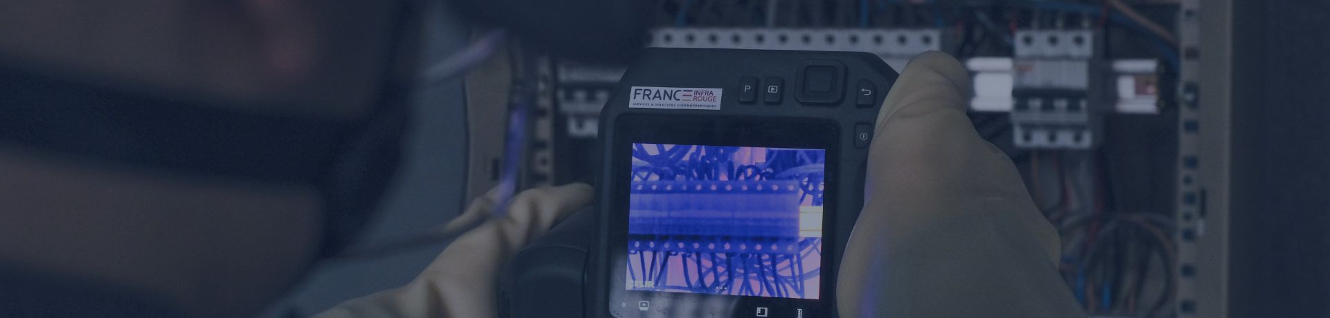 Un technicien de dos, ganté, utilise une caméra thermique pour inspecter un tableau électrique. L'écran de la caméra montre une image en couleurs vives des composants internes.