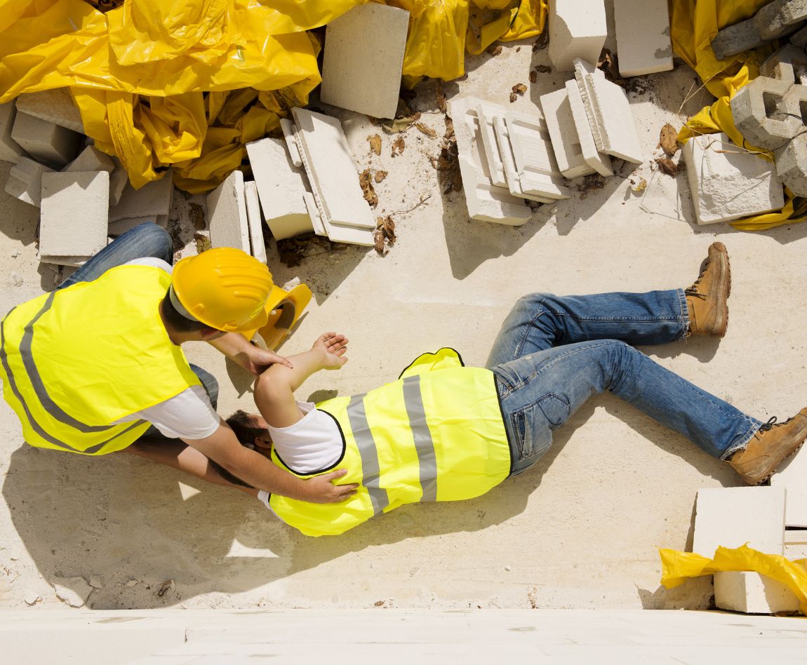 Vue en plongée d'un ouvrier blessé en gilet de sécurité jaune allongé au sol, secouru par un autre ouvrier.