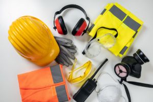 Une collection d'équipements de protection individuelle (EPI) : casque de sécurité jaune, gants de travail, lunettes de protection, casque anti-bruit, gilet haute visibilité et masque respiratoire, posés sur une surface blanche.