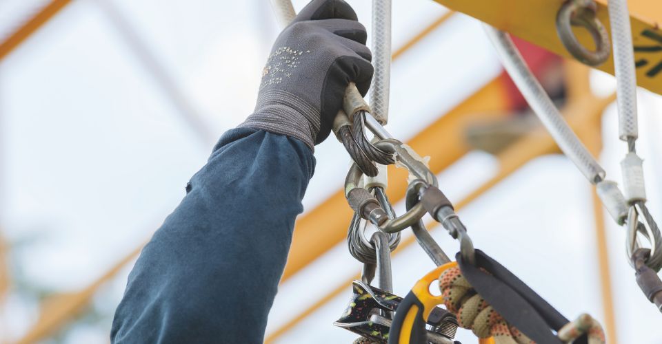 Main gantée d'un ouvrier accrochant des mousquetons de sécurité sur une structure jaune.