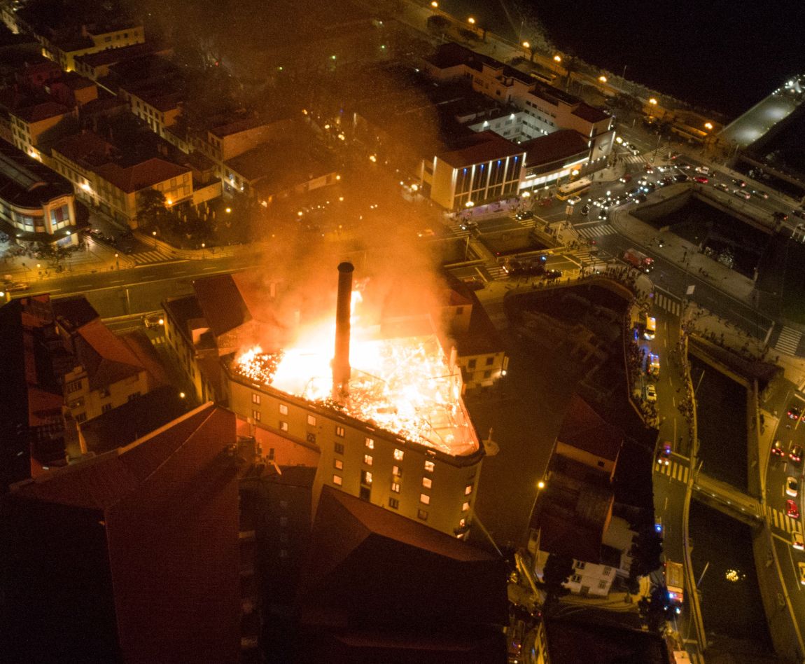 Vue aérienne nocturne d'un bâtiment industriel en flammes au cœur d'une zone urbaine, illustrant l'ampleur et la dangerosité d'un sinistre incendie majeur.
