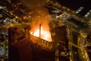 Vue aérienne nocturne d'un bâtiment industriel en flammes au cœur d'une zone urbaine, illustrant l'ampleur et la dangerosité d'un sinistre incendie majeur.