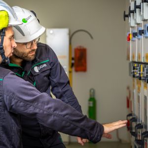 Deux techniciens en combinaison de travail noire et verte, portant des casques de chantier, examinent tableau électrique. L'un d'eux pointe du doigt l'équipement.