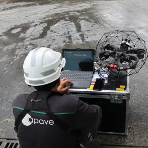 Un technicien en casque de chantier, accroupi, utilise un ordinateur portable à côté d'un drone sphérique posé sur une valise de transport.