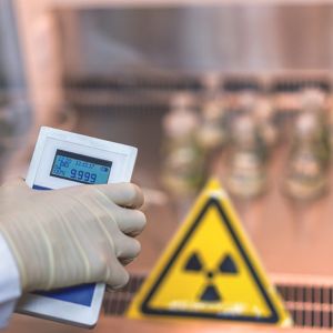 Une main gantée tenant un dosimètre portatif, affichant un taux de radiation, avec un panneau de signalisation triangulaire jaune indiquant un danger de radiation en arrière-plan.