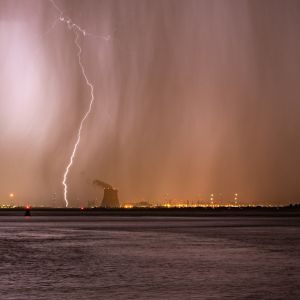 Un éclair frappe le ciel de nuit au-dessus de la silhouette sombre d'une tour de refroidissement de centrale nucléaire, avec un aperçu de lumières de ville au loin.