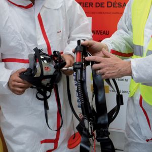 Deux techniciens en combinaison de travail blanche manipulent de l'équipement de sécurité et de protection respiratoire dans un environnement industriel amiante.