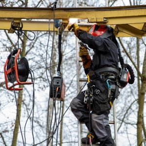 Un travailleur portant un masque de soudure et un harnais de sécurité est en train de souder une structure métallique en hauteur.