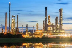 A night view of an illuminated oil refinery or petrochemical complex, with its chimneys and towers reflecting in the water.