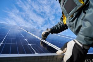 A technician wearing work gloves is inspecting and cleaning a row of solar panels against a blue sky with clouds.