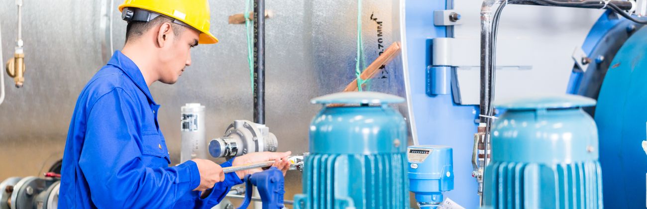 Un ouvrier en salopette bleue et casque de sécurité jaune, inspectant des machines industrielles avec des moteurs bleus.