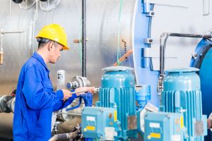 Un ouvrier en salopette bleue et casque de sécurité jaune, inspectant des machines industrielles avec des moteurs bleus.